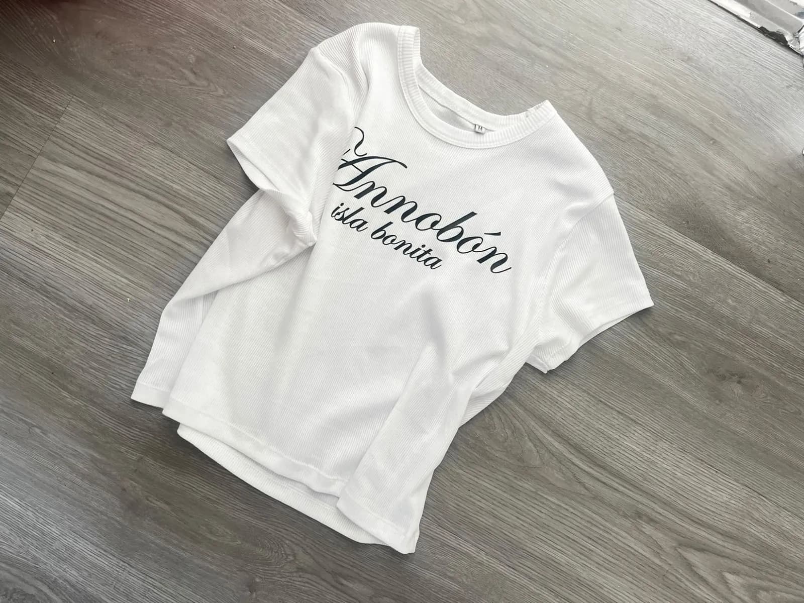 La Annobón Baby Tee extendida sobre un suelo de madera gris